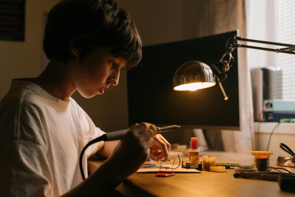Young teen focused on soldering circuit board at desk. Engaging in hands-on technical skills.